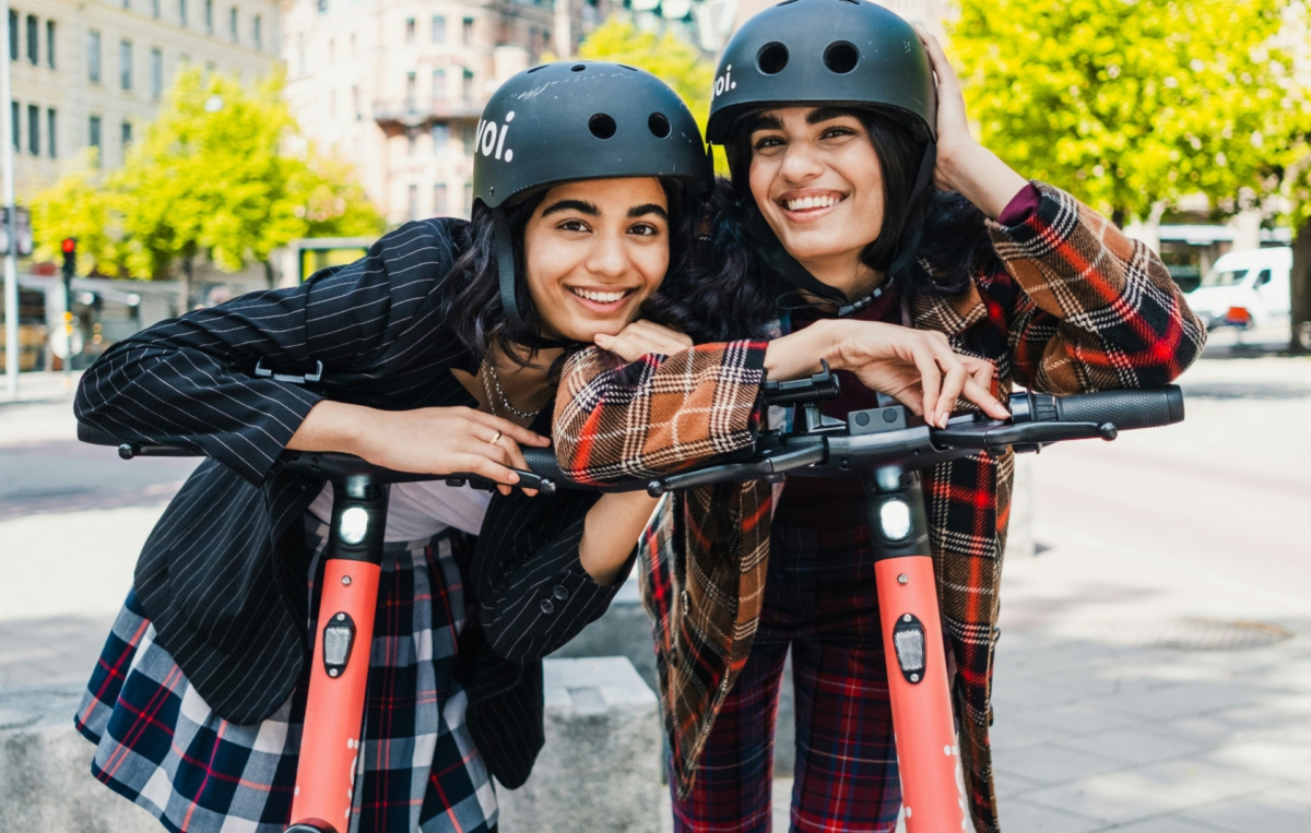 Zwei Frauen tragen Helme und lehnen sich auf einem E-Scooter an.