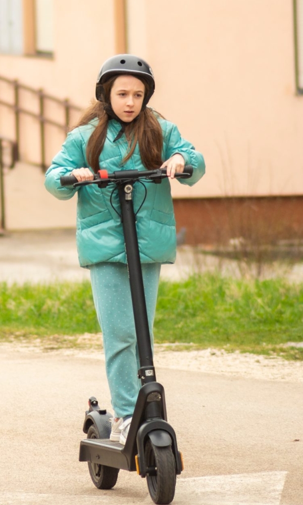 Ein Kind fährt mit einem E-Scooter auf der Straße, trägt einen Helm und ist in einer Wohngegend unterwegs.