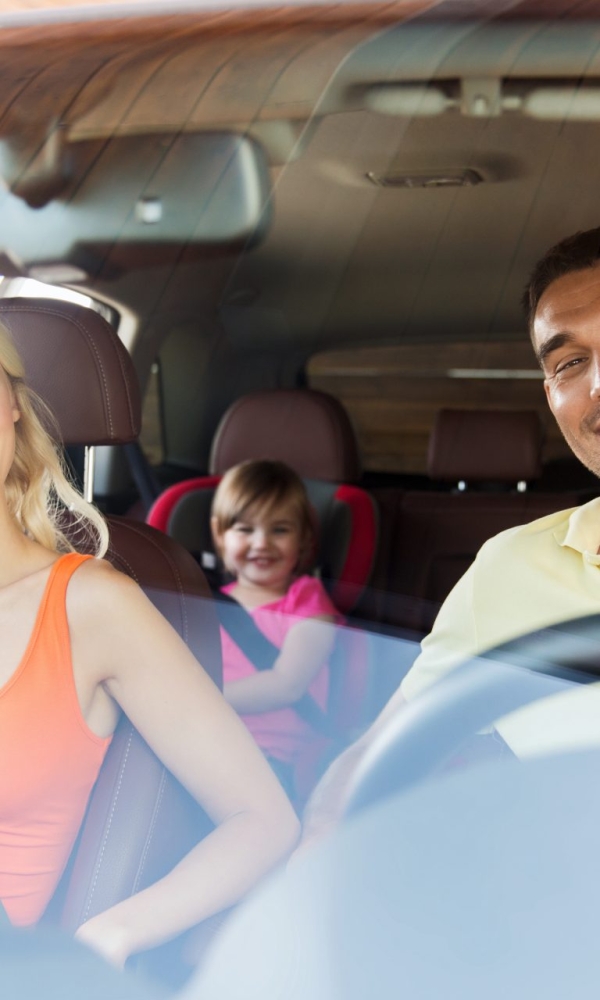 Ein Mann und eine Frau sitzen lachend auf den Vordersitzen eines Autos, während ein Kind auf der Rückbank im Kindersitz mitfährt und ebenfalls lächelt.