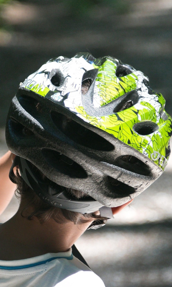Ein Kind von hinten, das auf einem Waldweg steht. Es trägt ein weißes T-Shirt und einen Fahrradhelm in Schwarz und Grün, auf den Sonnenlicht durch die Bäume fällt.