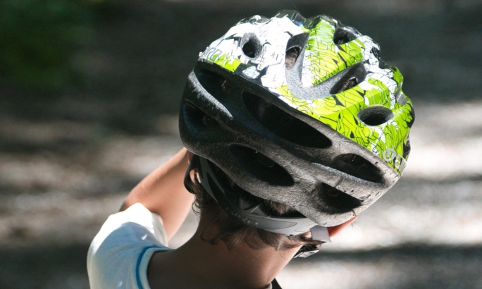 Ein Kind von hinten, das auf einem Waldweg steht. Es trägt ein weißes T-Shirt und einen Fahrradhelm in Schwarz und Grün, auf den Sonnenlicht durch die Bäume fällt.