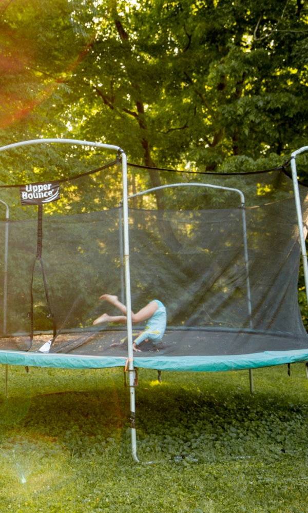 Eine Gartenszene bei Sonnenschein: Rechts steht ein großes Trampolin mit Sicherheitsnetz, auf dem ein Kind liegt. Links daneben spielt ein weiteres Kind mit einem Hund. Im Hintergrund sind dichte grüne Bäume und Sträucher zu sehen, durch die Sonnenstrahlen scheinen.