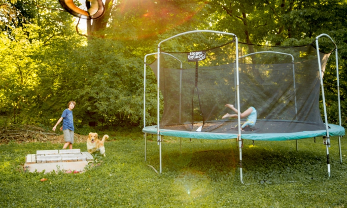 Eine Gartenszene bei Sonnenschein: Rechts steht ein großes Trampolin mit Sicherheitsnetz, auf dem ein Kind liegt. Links daneben spielt ein weiteres Kind mit einem Hund. Im Hintergrund sind dichte grüne Bäume und Sträucher zu sehen, durch die Sonnenstrahlen scheinen.