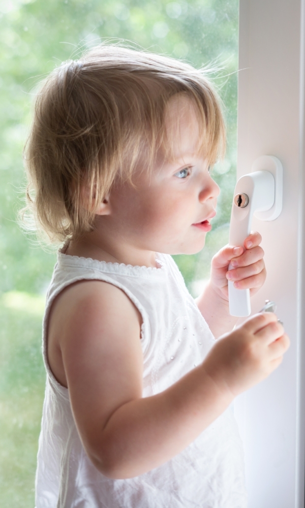 Ein kleines Kind steht an einem geschlossenen Fenster und hält die beiden weißen Fenstergriffe fest. Es trägt ein ärmelloses weißes Oberteil und blickt konzentriert nach draußen. Im Hintergrund sind unscharf grüne Bäume zu erkennen.