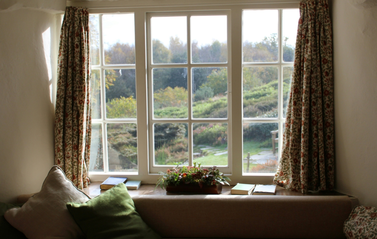 Ein großes Fenster mit weißen Sprossen gibt den Blick auf eine grüne Landschaft mit Bäumen und Wiesen frei. Links und rechts hängen geblümte Vorhänge. Vor dem Fenster steht eine breite Fensterbank mit Kissen, einem kleinen Pflanzkorb und mehreren Büchern.