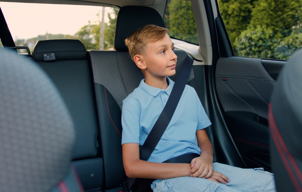 Ein Junge sitzt angeschnallt auf dem Rücksitz eines Autos. Er trägt ein hellblaues Poloshirt und blickt aus dem Fenster. Im Hintergrund sind Bäume und eine Straße zu erkennen.