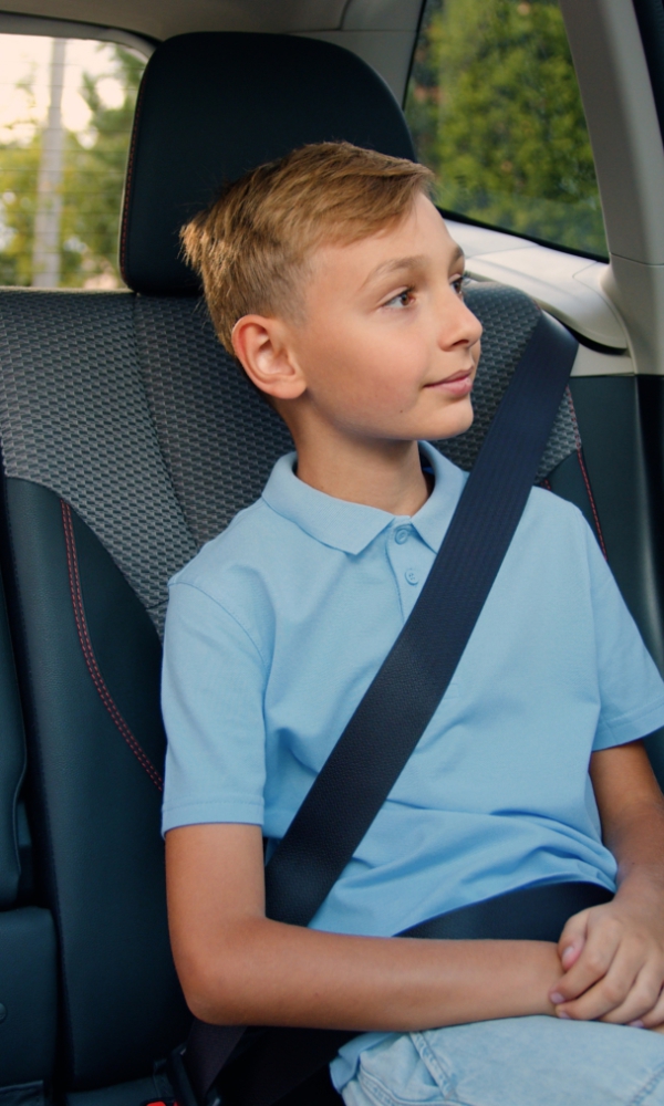 Ein Junge sitzt angeschnallt auf dem Rücksitz eines Autos. Er trägt ein hellblaues Poloshirt und blickt aus dem Fenster. Im Hintergrund sind Bäume und eine Straße zu erkennen.
