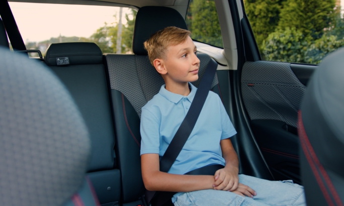 Ein Junge sitzt angeschnallt auf dem Rücksitz eines Autos. Er trägt ein hellblaues Poloshirt und blickt aus dem Fenster. Im Hintergrund sind Bäume und eine Straße zu erkennen.