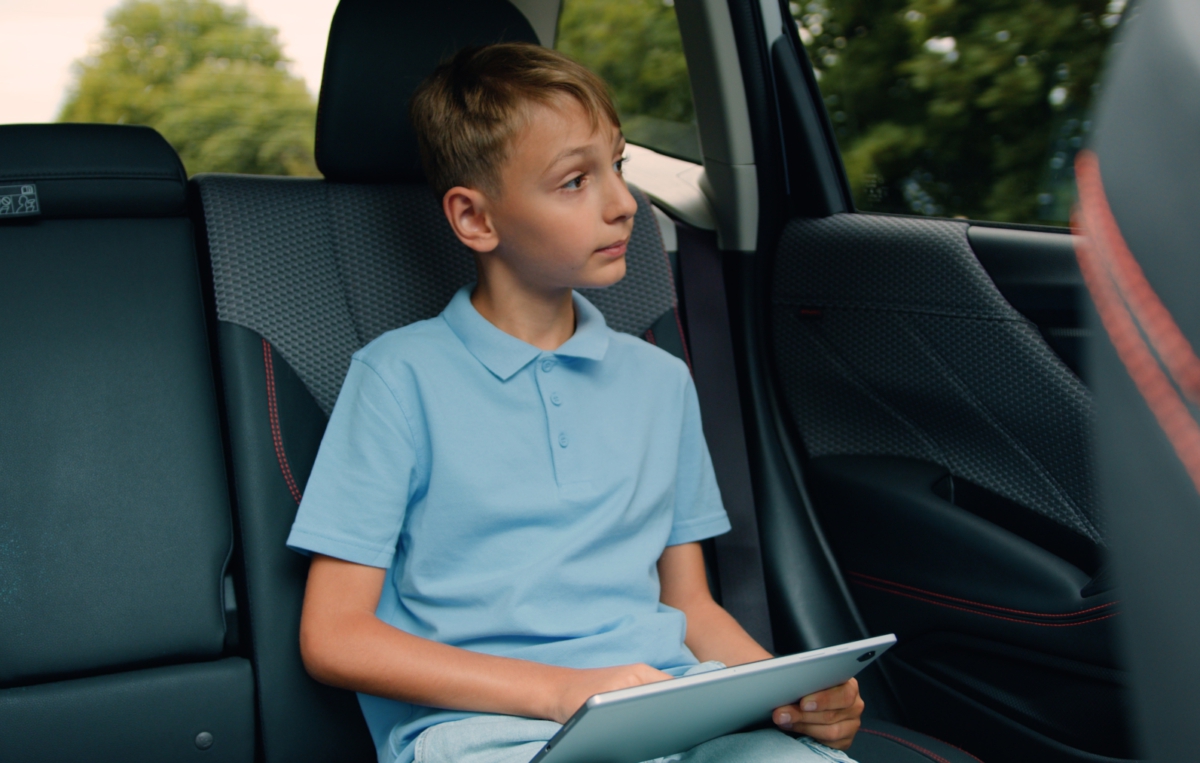 Ein Junge sitzt auf dem Rücksitz eines Autos und hält ein Tablet in den Händen. Er trägt ein hellblaues Poloshirt und blickt seitlich aus dem Fenster. Im Hintergrund sind verschwommene Bäume zu erkenne