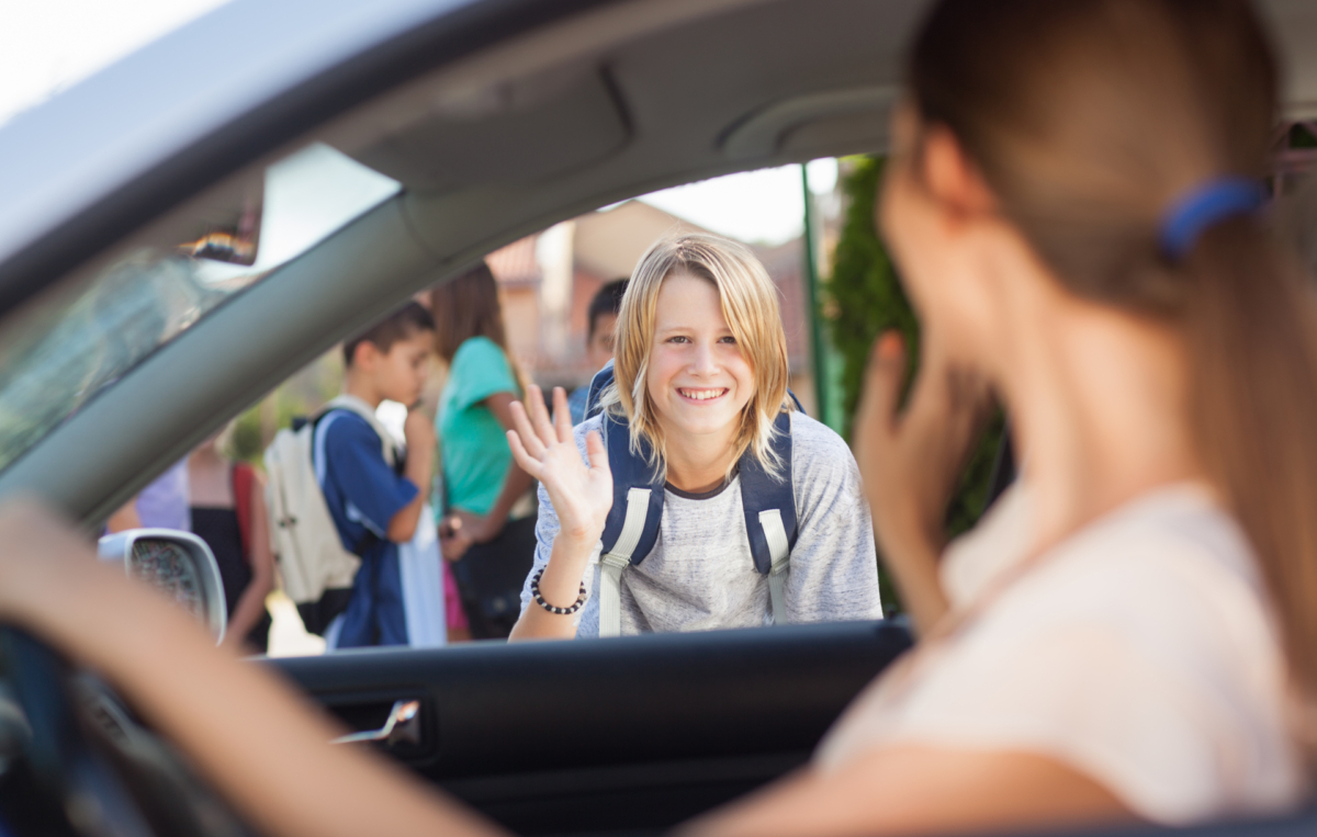 Schüler mit Rucksack winkt lachend einer Frau im Auto zum Abschied, andere Kinder stehen im Hintergrund.