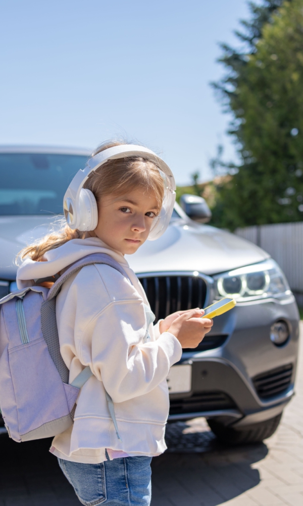 Mädchen mit Kopfhörern und Rucksack steht mit einem Smartphone in der Hand vor einem Auto auf der Straße.