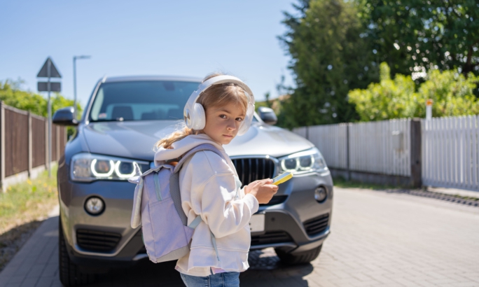Mädchen mit Kopfhörern und Rucksack steht mit einem Smartphone in der Hand vor einem Auto auf der Straße.