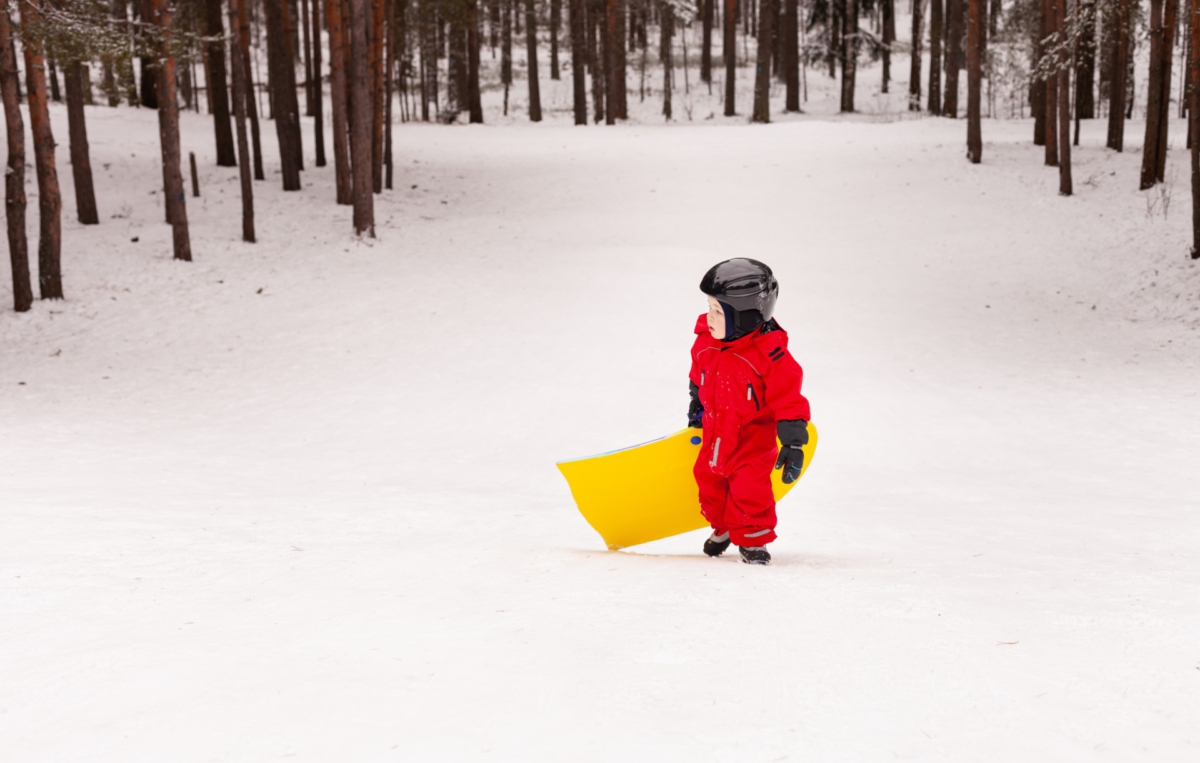 Ein Kind in einem roten Schneeanzug mit schwarzem Helm steht im Schnee und hält einen großen gelben Kunststoff-Schlitten in der Hand. Der Hintergrund zeigt eine verschneite Waldlichtung mit vielen schlanken Bäumen.