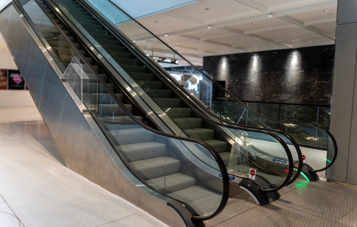 Leere Rolltreppe in einem modernen Einkaufszentrum oder Gebäude, mit Glasgeländern und metallischen Seitenwänden.