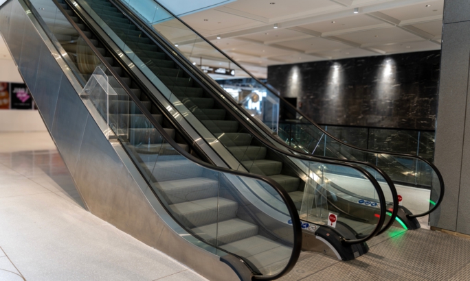 Leere Rolltreppe in einem modernen Einkaufszentrum oder Gebäude, mit Glasgeländern und metallischen Seitenwänden.
