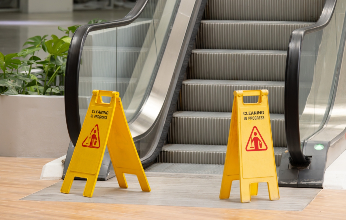 Gelbe Warnschilder mit der Aufschrift „Caution – Wet Floor“ stehen vor einer stillstehenden Rolltreppe in einem Einkaufszentrum.