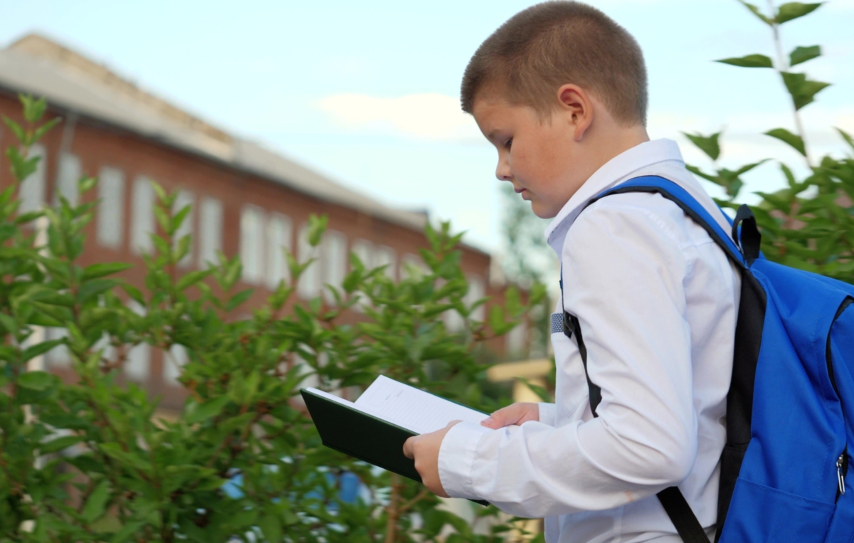 Ein Schuljunge mit weißem Hemd und blauem Rucksack liest im Freien in einem Buch.