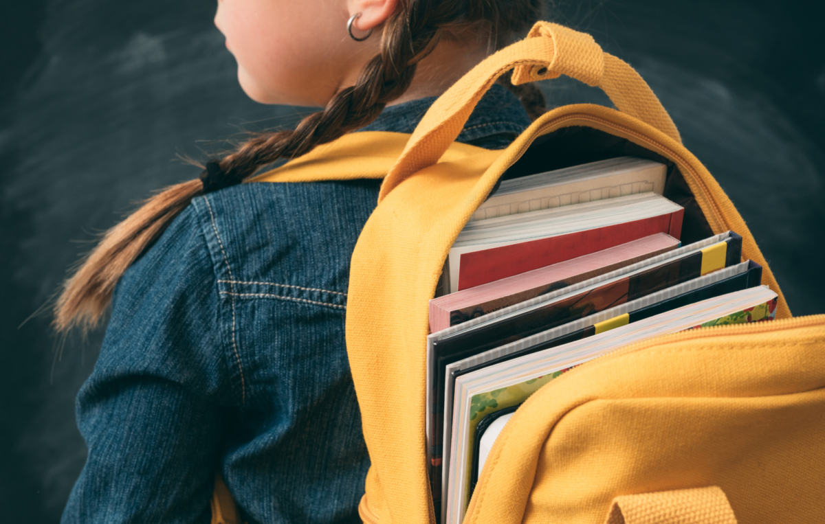 Ein Schulmädchen mit zwei Zöpfen trägt einen gelben Rucksack, der mit Büchern gefüllt ist.