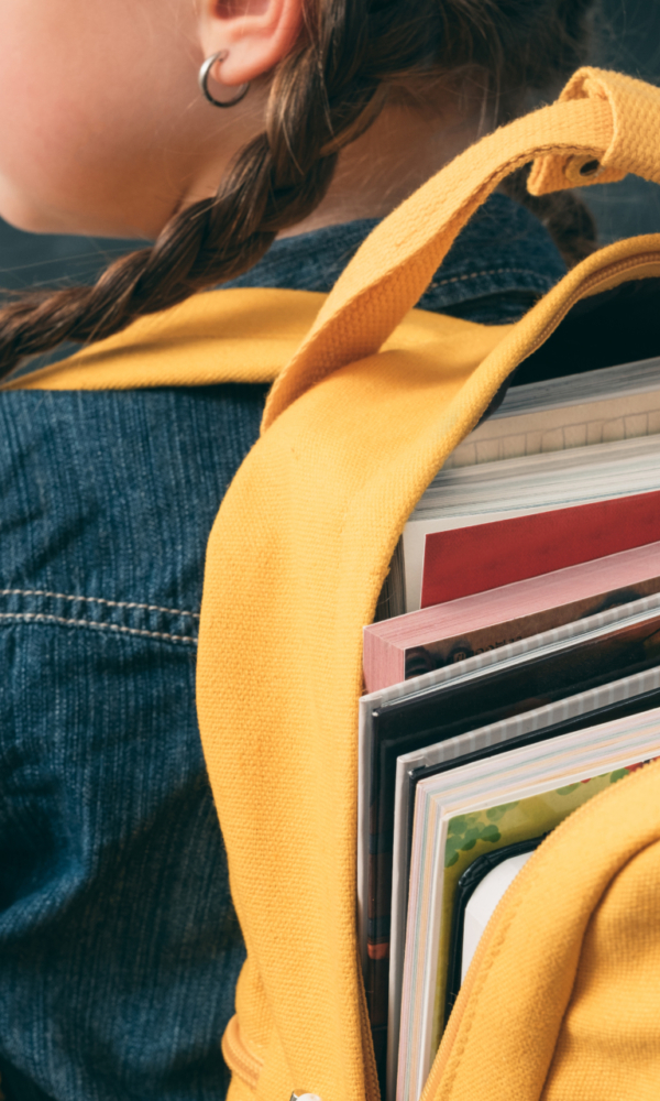 Ein Schulmädchen mit zwei Zöpfen trägt einen gelben Rucksack, der mit Büchern gefüllt ist.