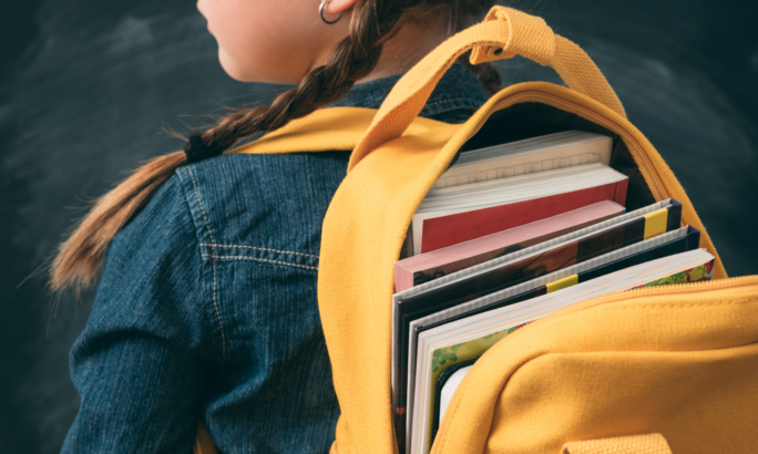 Ein Schulmädchen mit zwei Zöpfen trägt einen gelben Rucksack, der mit Büchern gefüllt ist.