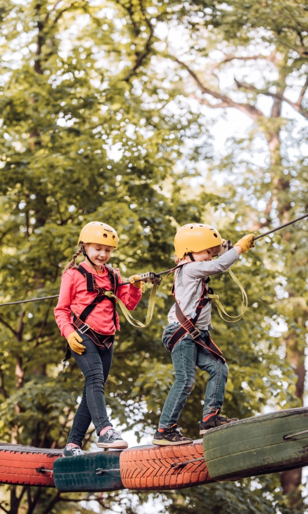 Zwei Kinder klettern im Hochseilgarten. Beide tragen Schutzhelme, Sicherheitsgurte und sind mit Seilen gesichert. Das linke Kind trägt eine rote Jacke, das rechte Kind ein graues Oberteil. Sie bewegen sich konzentriert über eine erhöhte Plattform vor einem Hintergrund mit grünen Bäumen.