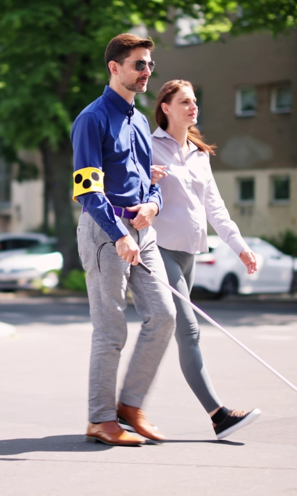 Ein blinder Mann mit Blindenstock und Armbinde überquert gemeinsam mit einer Begleiterin eine Straße.