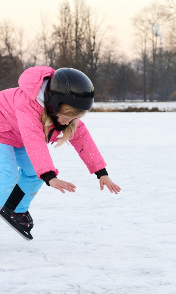 Ein Kind läuft mit Schlittschuhen über eine Eisfläche. Es trägt eine pinkfarbene Jacke, eine hellblaue Hose, Handschuhe und einen schwarzen Helm. Das Kind beugt sich nach vorne und streckt die Arme aus, als würde es das Gleichgewicht halten oder gerade hinfallen. Im Hintergrund sind kahle Bäume im Winter zu sehen.