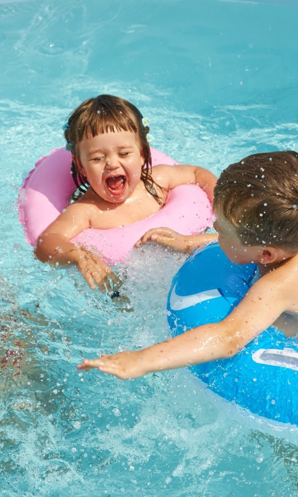 Drei Kinder spielen fröhlich im Wasser eines Pools. Zwei von ihnen sitzen in bunten Schwimmringen (rosa und blau), während das dritte Kind neben ihnen im Wasser planscht. Alle lachen und spritzen Wasser, es herrscht ausgelassene Sommerstimmung.