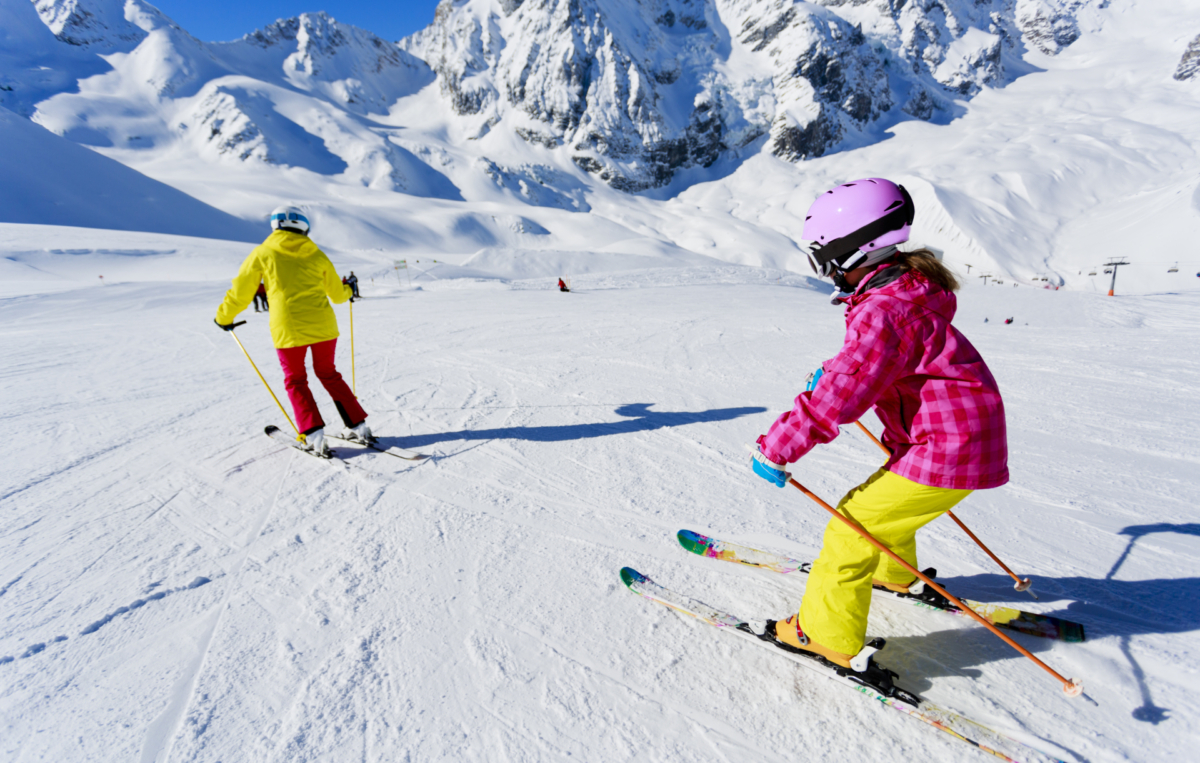 Zwei Kinder fahren bei sonnigem Wetter auf einer verschneiten Piste Ski. Im Hintergrund sind hohe, schneebedeckte Berge zu sehen.