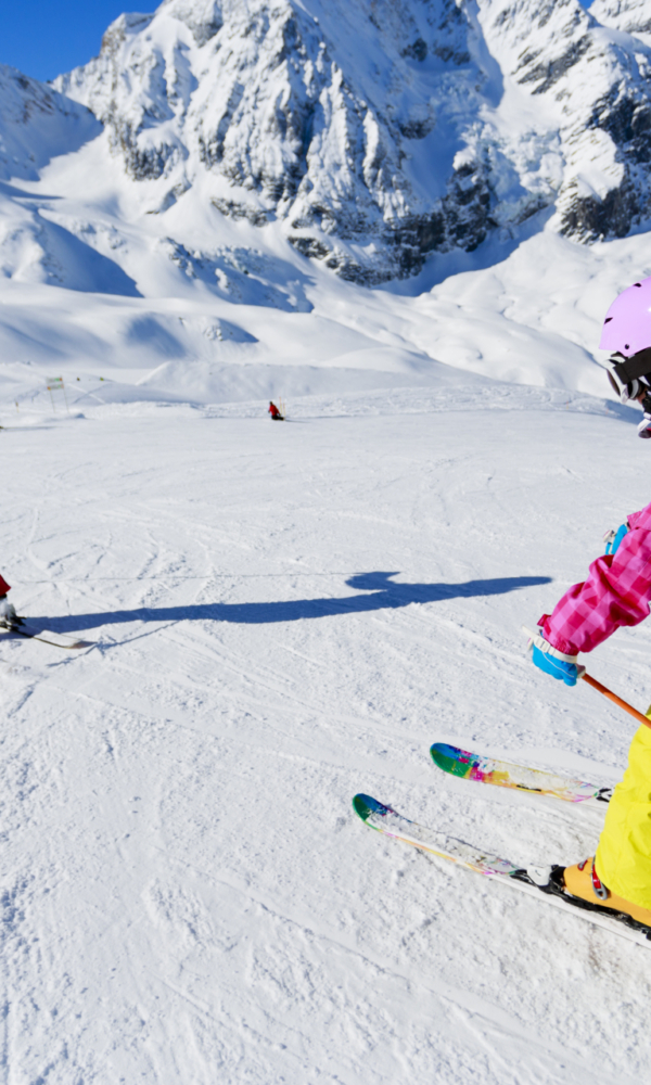 Zwei Kinder fahren bei sonnigem Wetter auf einer verschneiten Piste Ski. Im Hintergrund sind hohe, schneebedeckte Berge zu sehen.