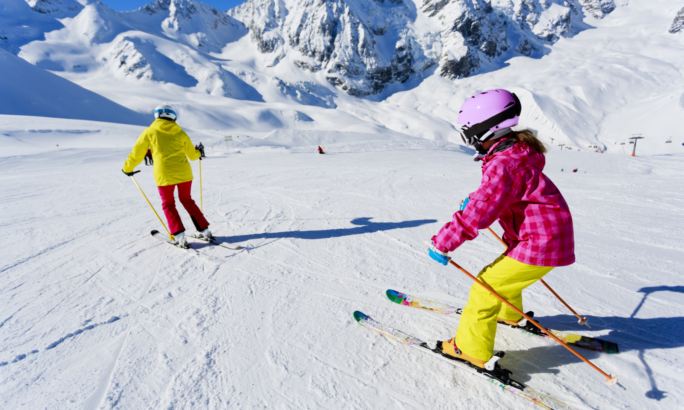 Zwei Kinder fahren bei sonnigem Wetter auf einer verschneiten Piste Ski. Im Hintergrund sind hohe, schneebedeckte Berge zu sehen.