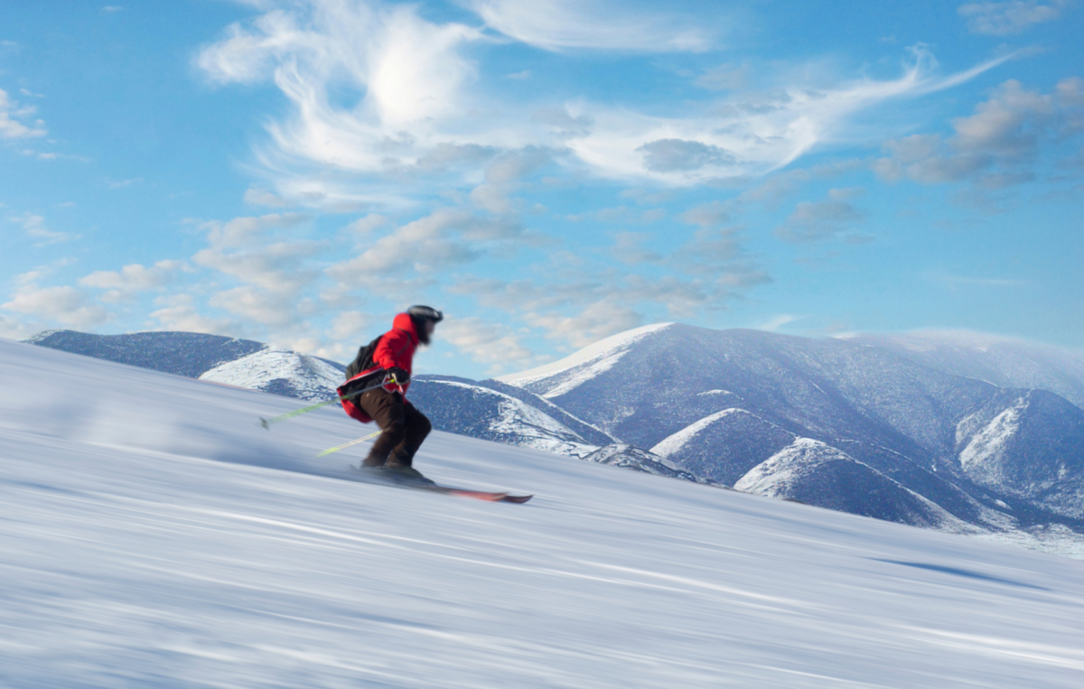 Eine Person fährt mit Skiern einen verschneiten Hang hinunter. Im Hintergrund sind schneebedeckte Berge und ein blauer Himmel mit Wolken zu sehen.