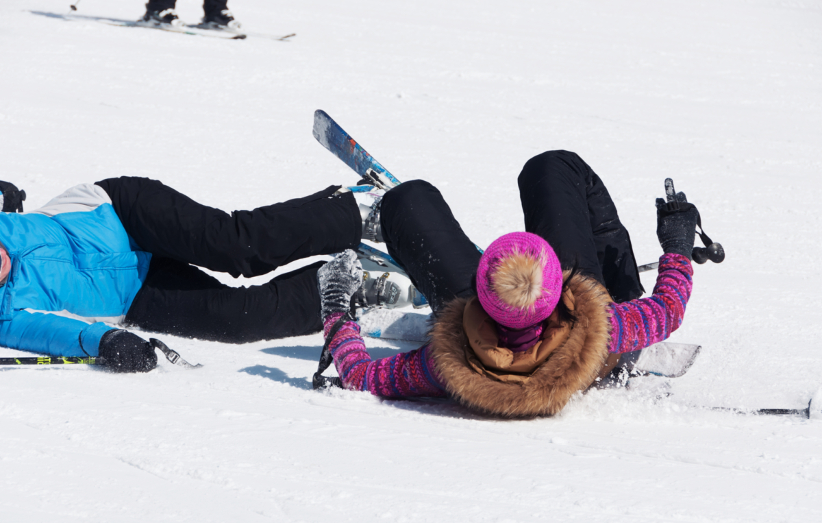 Zwei Personen liegen nach einem Sturz auf der Skipiste im Schnee. Beide tragen Ski und Winterkleidung.