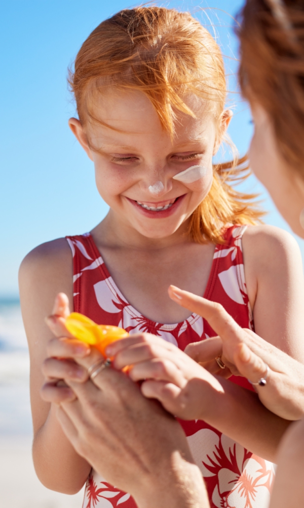 Ein Kind im Badeanzug am Strand lächelt, während eine erwachsene Person Sonnenschutzcreme auf ihre Hände gibt. Im Hintergrund ist das Meer zu sehen.