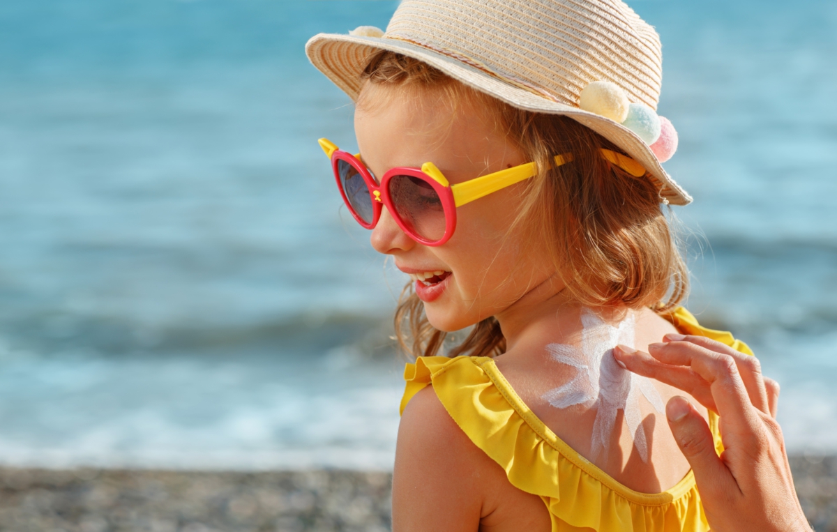 Ein Kind mit Sonnenhut und roter Sonnenbrille sitzt am Strand. Es trägt ein gelbes Kleid mit Rüschen und lacht, während ihm Sonnencreme auf den Rücken aufgetragen wird. Im Hintergrund ist das Meer zu sehen.