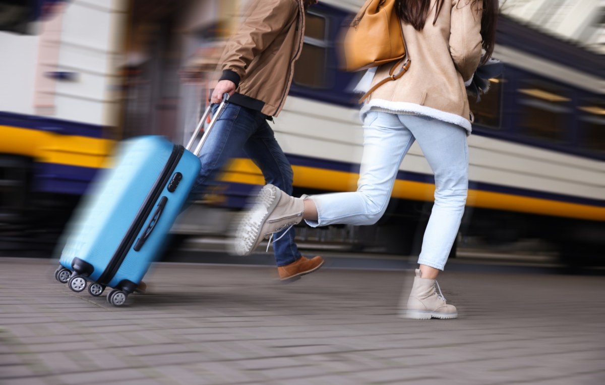 Zwei Personen laufen mit einem blauen Rollkoffer über den Bahnsteig, während im Hintergrund ein Zug steht.