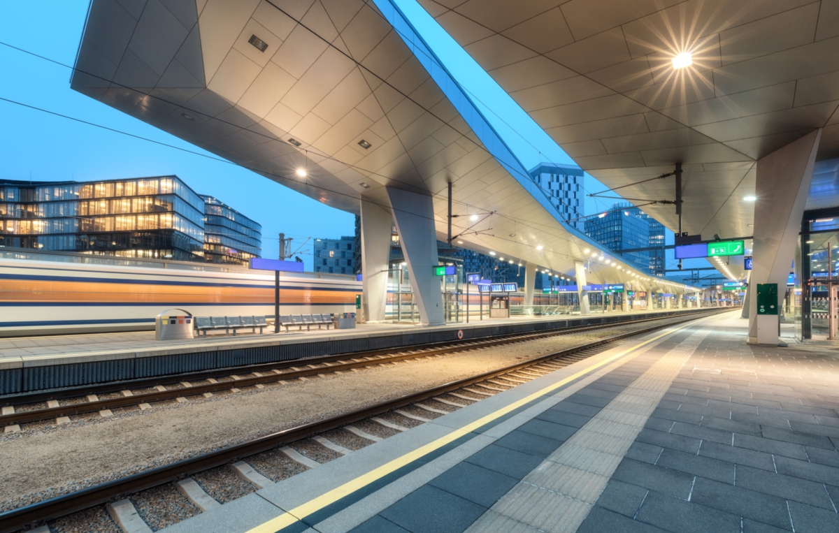 Moderner, beleuchteter Bahnhof am Abend mit Bahnsteigen, Gleisen und vorbeifahrendem Zug, umgeben von städtischen Gebäuden.