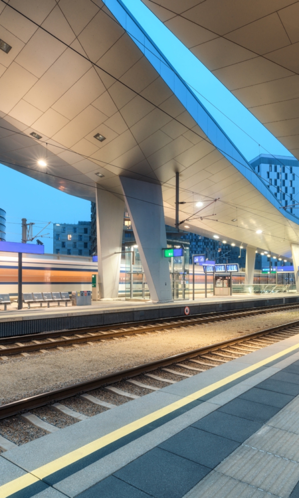 Moderner, beleuchteter Bahnhof am Abend mit Bahnsteigen, Gleisen und vorbeifahrendem Zug, umgeben von städtischen Gebäuden.