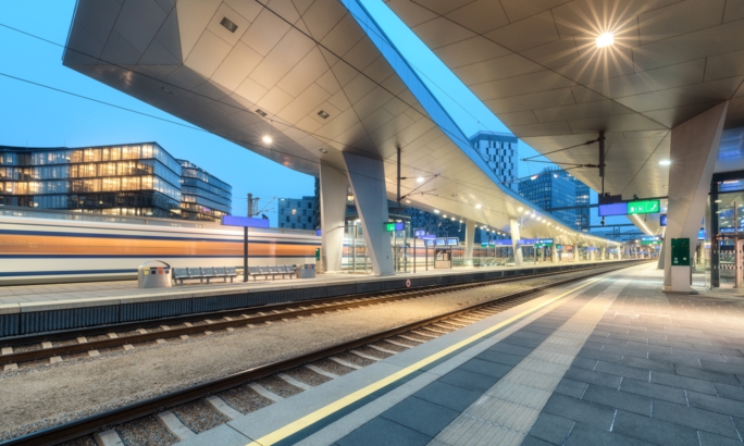 Moderner, beleuchteter Bahnhof am Abend mit Bahnsteigen, Gleisen und vorbeifahrendem Zug, umgeben von städtischen Gebäuden.