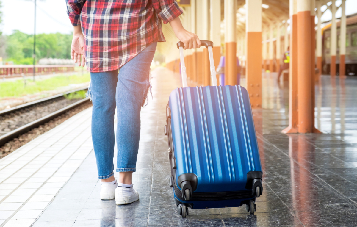 Person zieht einen blauen Rollkoffer über den Bahnsteig eines Bahnhofs, im Hintergrund sind Gleise und Säulen zu sehen.