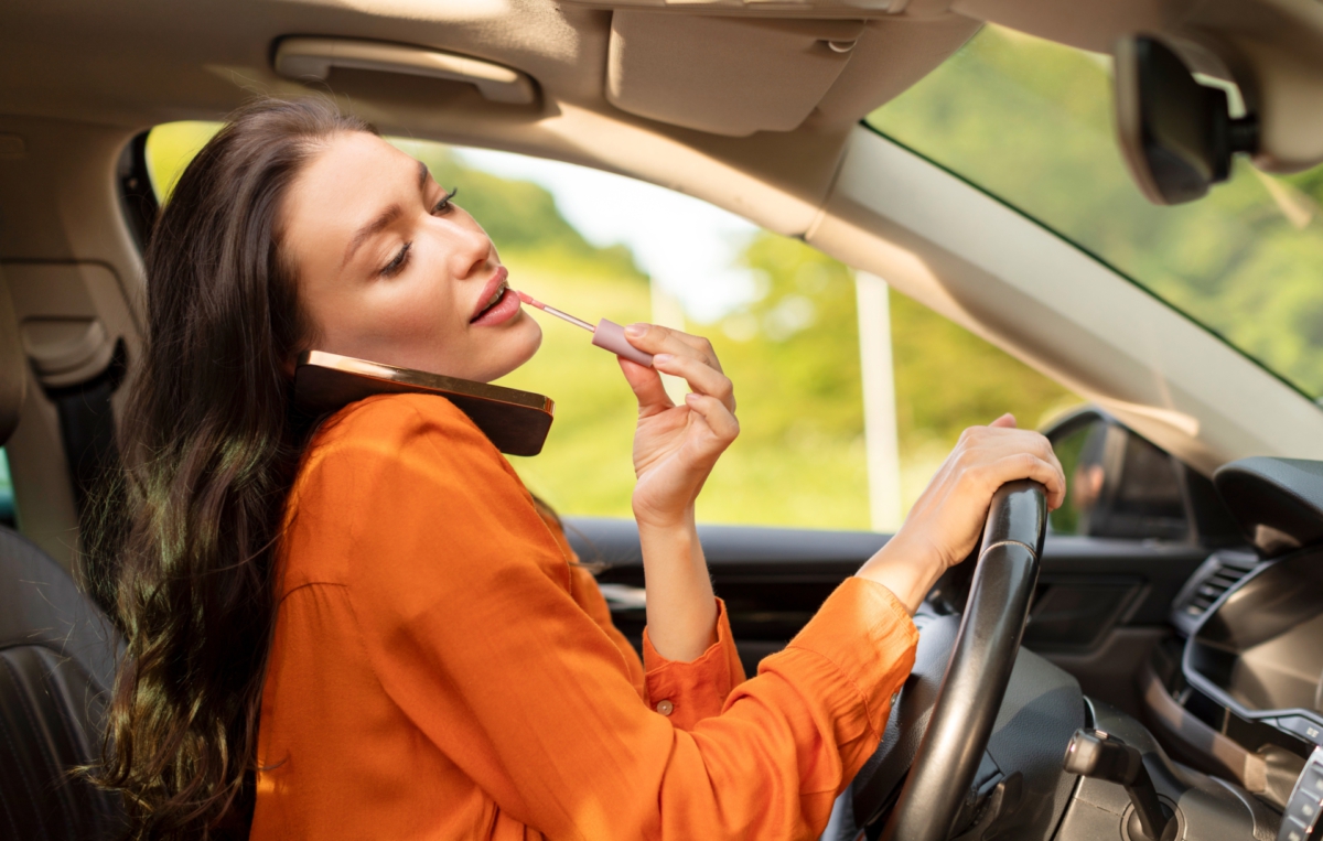 Eine Frau sitzt im Auto am Steuer, telefoniert und schminkt sich gleichzeitig.