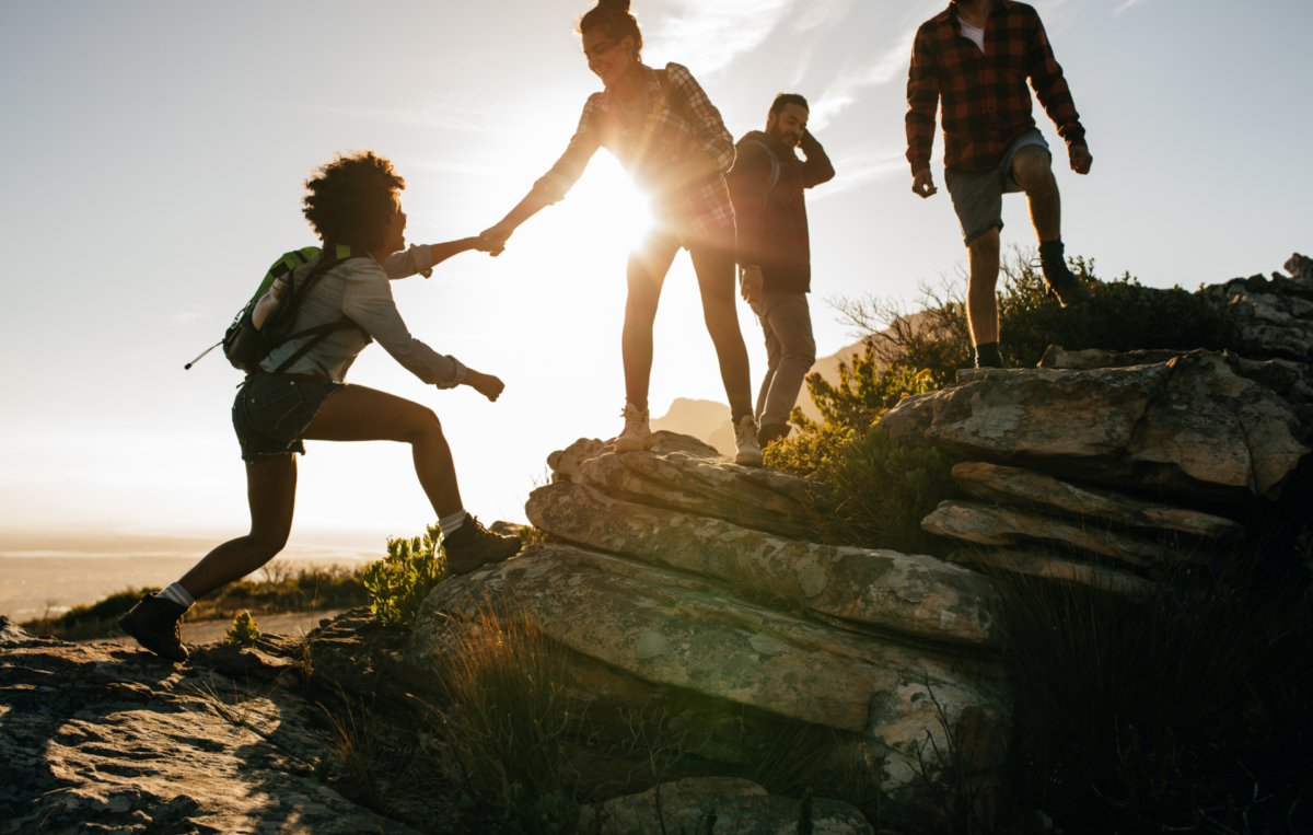Gruppe von Freunden wandert in den Bergen, eine Person hilft der anderen beim Aufstieg über Felsen, im Hintergrund scheint die Sonne.