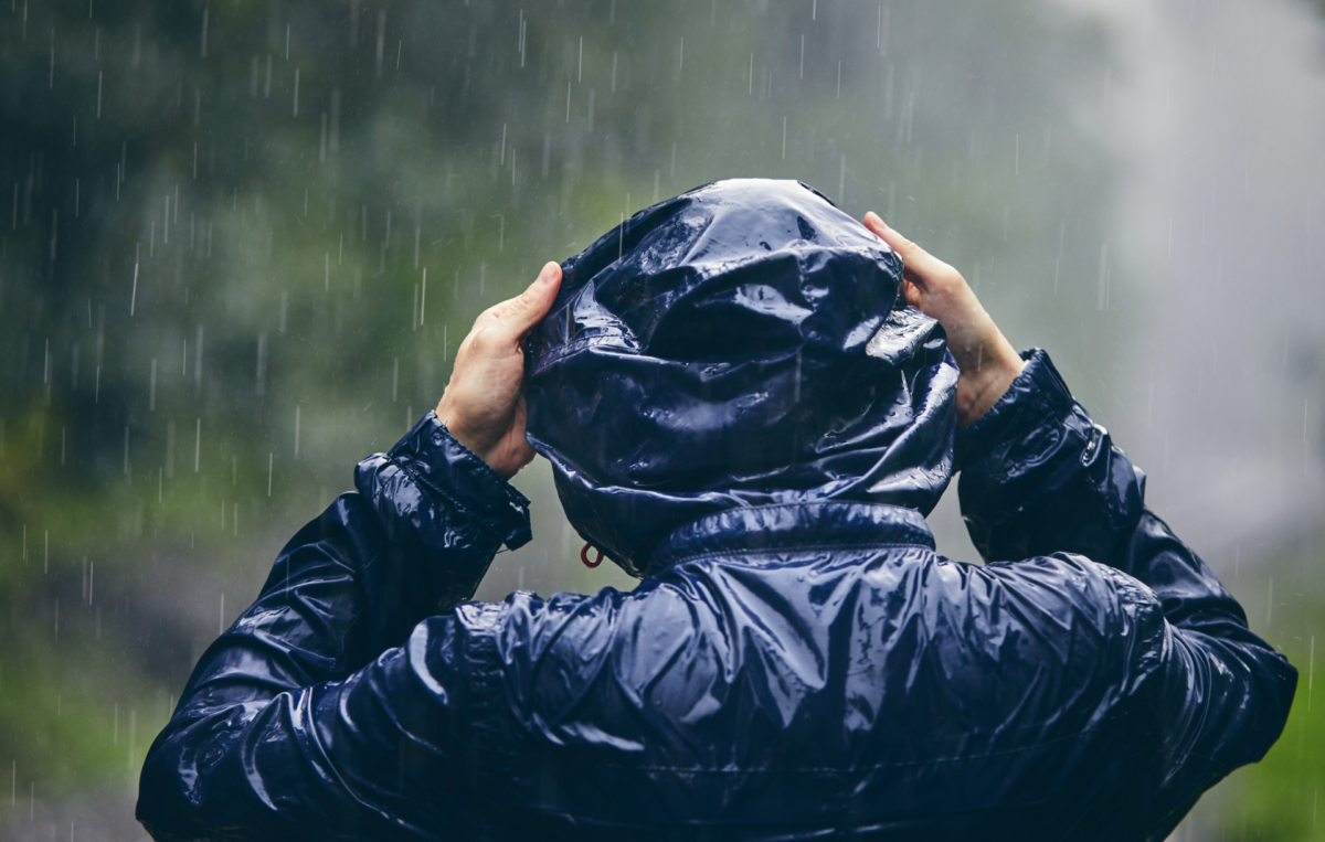 Person in dunkelblauer Regenjacke zieht sich die Kapuze über den Kopf, während starker Regen fällt.