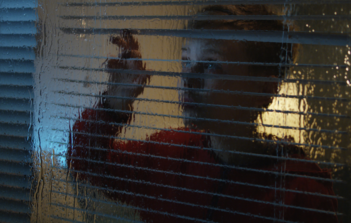 Kind in rotem Pullover blickt nachdenklich durch eine verregnete Fensterscheibe und legt die Hand an das Glas.