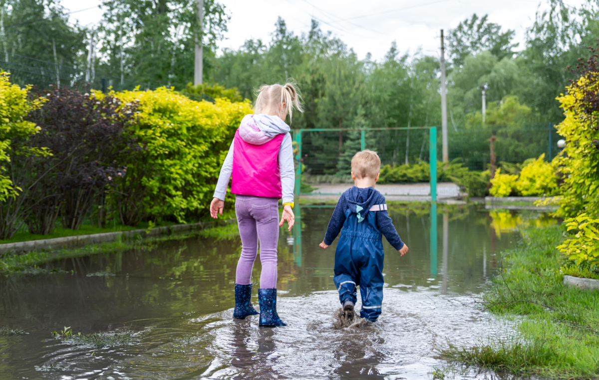 Zwei Kinder spielen in Regenstiefeln auf einer überfluteten Straße, umgeben von Bäumen und Sträuchern.