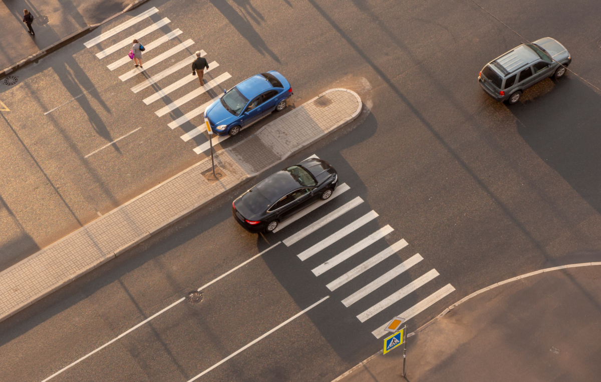 Straßenkreuzung mit Zebrastreifen und Verkehrsinsel, darauf warten Fußgänger; Autos fahren auf der Straße, Ansicht aus der Vogelperspektive.