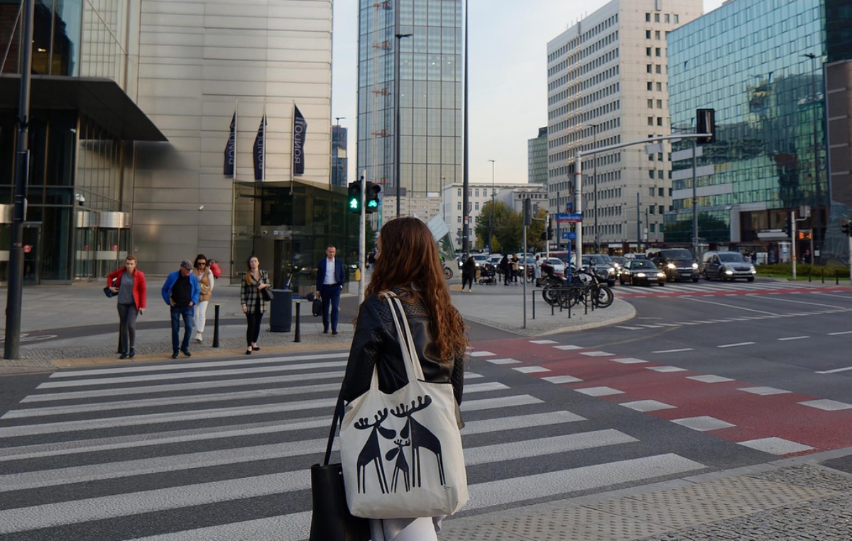 Frau mit Tasche überquert einen Zebrastreifen in einer modernen Innenstadt mit Hochhäusern und Glasfassaden..