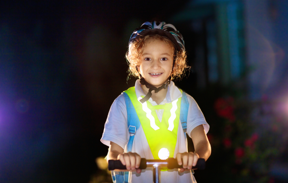 Ein lachendes Kind fährt bei Dunkelheit auf einem Roller. Es trägt einen Fahrradhelm, einen Rucksack und eine reflektierende Warnweste, die im Licht hell leuchtet. Vorne am Roller ist ein Licht angebracht, das die Szene zusätzlich erhellt.