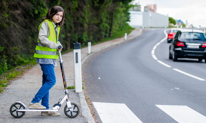 Ein Kind steht mit einem Tretroller am Straßenrand neben einem Zebrastreifen. Es trägt eine reflektierende Warnweste und schaut nach vorne. Auf der Straße fahren mehrere Autos vorbei. Rechts ist eine leicht kurvige Fahrbahn mit Leitpfosten, links ein Grünstreifen mit Bäumen.