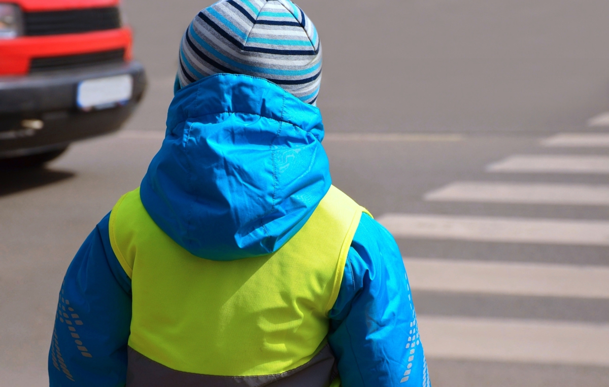 Ein Kind steht vor einem Zebrastreifen. Es trägt eine blau-türkise Winterjacke, eine reflektierende Sicherheitsweste und eine gestreifte Wollmütze. Der Blick ist von hinten aufgenommen, sodass das Kind in Richtung Straße schaut. Auf der Straße ist ein vorbeifahrendes Fahrzeug zu erkennen